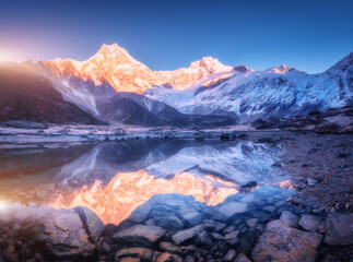Wall Mural - Snowy mountain with illuminated peaks is reflected in beautiful lake at sunrise. Landscape with lighted rocks, violet sky, pond, stones in water at dawn. Winter Himalayan mountains in Nepal. Nature
