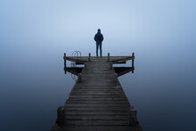 A Man Looking Over A Lake From A Jetty On A Foggy, Autumn Day.