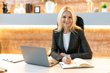 Wall Mural - Confident intelligent caucasian middle aged business woman, financial manager, broker, in formal suit, sitting at table in office, using laptop, taking notes, looking at camera, smiling friendly