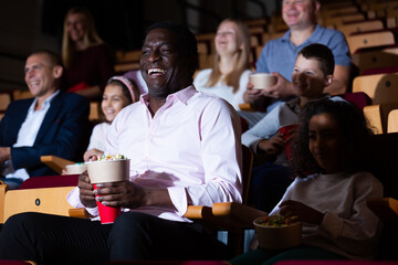 Wall Mural - Portrait of cheerful laughing african american man sitting in movie theater with popcorn and watching comedy..