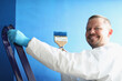 © H_Ko - Painter holding brush covered in blue colour for painting walls, worker in uniform