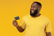 © ViDi Studio - Young smiling satisfied fun rich cool happy black man 20s wearing bright casual t-shirt hold in hand credit bank card look aside on workspace isolated on plain yellow color background studio portrait.
