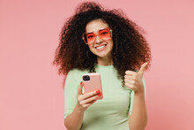 Fun Young Curly Latin Woman 20s Years Old Wears Mint T-shirt Sunglasses Hold In Hand Use Mobile Cell Phone Showing Thumb Up Like Gesture Isolated On Plain Pastel Light Pink Background Studio Portrait.