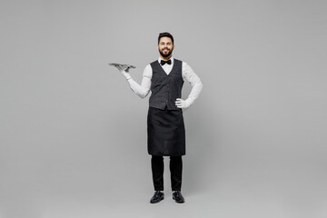 Wall Mural - Full body young fun barista male waiter butler man wear white shirt vest elegant uniform work at cafe hold carry metal tray stand akimbo isolated on plain grey background Restaurant employee concept.