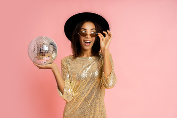 Wall Mural - Celebrating woman in golden sequins dress  posing with disco baloon over pink bacground in studio.