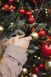 © Dina - woman holding mobile phone standing near christmas tree, texting or using social media