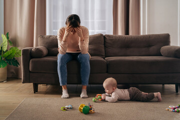 Wall Mural - Young single frustrated woman hold her head with hands sitting on sofa in living room while her toddler son playing with toys on floor. Exhausted mom suffering from postpartum post-natal depression 