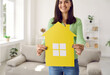 © Studio Romantic - Close up of yellow paper mock up of house in hands of woman who is happy owner of new home. Woman standing in living room shows mock up to camera. Concept of buying your own home. Selective focus.