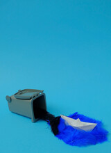 Spilled Black And Blue Feathers From A Trash Can And A White Paper Boat On A Light Blue Background. Text Space. Water Pollution, Waste Water, Creative, Concept. Minimal Style.
