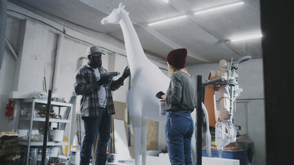 Woman mixing paint in cup and checking statue while black man drying white pigment on giraffe near robotic arm milling machine during work