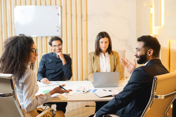 Business discussion in contemporary office of multiracial team. They are sitting around the table and listening each other with pleasure smiles