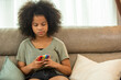 Depressed little African child girl sitting on sofa in living room with looking on mobile phone screen with sadness face. Social issue, family problem, cyberbullying and children mental health concept