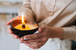 Close-up cropped shot of unrecognizable young woman holding burning scented handmade candle in hands. Closeup of female holding aromatic relaxing candle at home. Concept of wellness and relaxation.