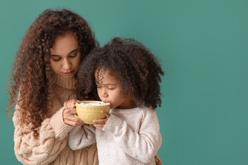 Poster - Little African-American girl and her mother with mug of hot cocoa on green background