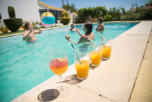 Close-up Of Glasses With Juice. Row Of Glasses With Bright Fresh Beverage Near Swimming Pool. People In Background. Party, Food, Drink Concept