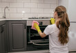 Young brunette girl in rubber yellow gloves rubs the stove oven with a rag in a gray and white kitchen