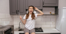 Young Brunette Girl In A Gray And White Kitchen Fooling Around Holding A Whisk In Her Right Hand And A Plate Over Her Head Instead Of A Hat