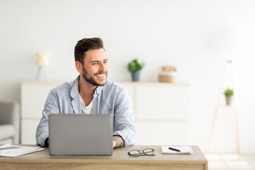 Happy freelance man working on laptop in home office, sitting on workplace and looking aside at free space