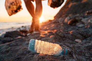 Plastic bottle lie at foreground. On the backgroud person holding two large bags of garbage in hands. Sunset on the background. Close up. The concept coastal zone cleaning