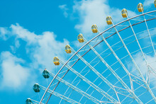 A Fragment Of A Ferris Wheel, Yellow And Green Cabins Against A Blue Sky With White Clouds