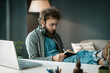 © Svyatoslav Lypynskyy - Student Listens to an Online Lecture and Makes Notes in a Notebook. a Man Writes Important Information in a Diary. Headphones Around his Neck. Feet on the Table. Close-up