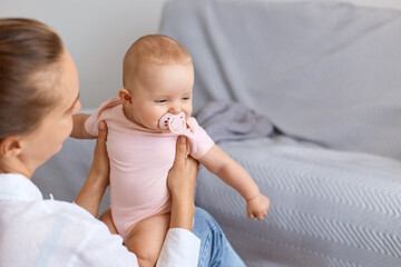 Wall Mural - Indoor shot of young adult female spending time with her toddler daughter with nipple, posing at home with gray sofa on background, happy childhood and parenthood.