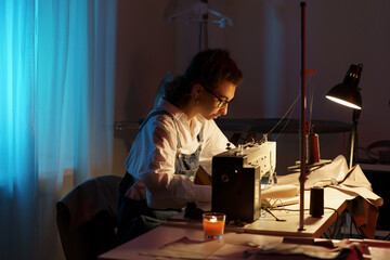 Wall Mural - Busy tailor woman working late at night sit at table with sewing machine, fabric and paper patterns in studio creating new collection for small atelier opening. Tailoring entrepreneurship concept