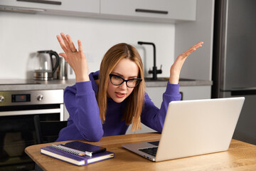Annoyed young adult woman in glasses work study at kitchen using laptop. Remote education