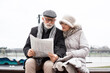 © Stock Rocket - An elderly couple sits on a bench at the waterfront in an urban area and reads the newspaper. The couple is smiling and looking happy in love.