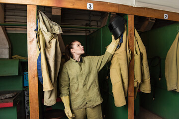 Wall Mural - Brunette welder in uniform taking protective mask in dressing room of factory