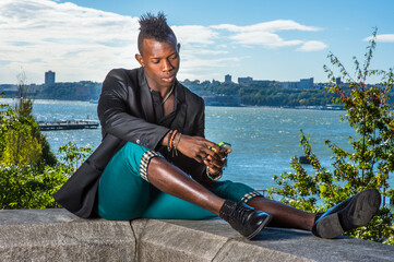 Wall Mural - Dressing in a black blazer, green pants and leather shoes, a young black guy with mohawk hair is sitting by a river, reading message on a mobile phone..