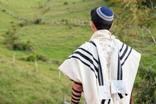 Jewish Man Praying With Talit, Kippah And Tefillin In Nature With Beautiful Green Meadow In The Background.