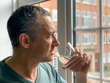 Man Is Drinking Water From Glass, Staying By The Window In Apartment. Middle Aged Man Is Drinking Water To Stay Hydrated Thirsty Man Having Hangover Pure Life Still Water