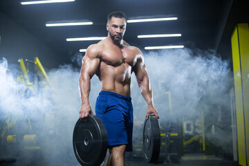 Handsome muscular athlete man is standing in a gym against the background of smoke holding barbell discs in both hands