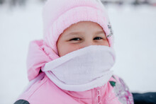 Close-up Photo Of Female Kid Looking Into Camera And Smiling Wearing White Scarf Covering Her Mouth And Pink Winter Hat And Pink Coat. Astonishing Background Full Of White Color And Snow. 