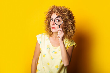 Wall Mural - curly young woman looks through a magnifying glass on a yellow background.