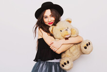 Close Up  Portrait Of Pretty Cute Young Woman In Stylish Hipster Outfit  With Big Eyes , Red Lips And Perfect Skin  Holding Her Fluffy Teddy Bear Against White Urban Wall.
