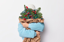 New Year And Christmas Concept. Unrecognizable Woman Hides Behind Big Fir Tree Bouquet Decorates With Garlands Awaits For Coming Holidays Dressed In Outerwear Poses Against White Background.