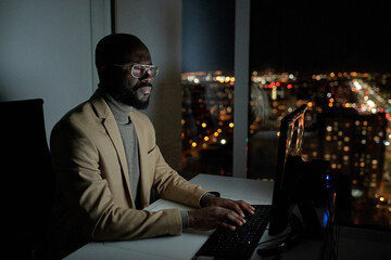 Poster - Bearded African office worker in formalwear typing and looking at computer screen while working overtime at night