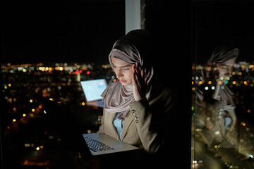 Poster - Young stressed Muslim businesswoman looking at laptop display and touching temple while standing by window