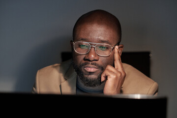 Poster - Serious young businessman in eyeglasses and formalwear sitting in front of computer monitor while working in office at night