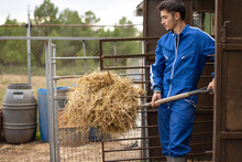 Portrait Of Young Farmer In Uniform Working On His Farm Clearing Straw From The Barn With A Sickle