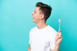 Young Brazilian man brushing teeth isolated on blue background laughing in lateral position