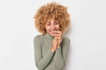 Wall Mural - Joyful young woman with pleasant appearance curly hair laughs out happily shows white teeth hears something funny wears casual jumper poses indoor. Real people emotions and happiness concept