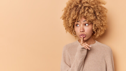 Wall Mural - Keep mouth shut. Mysterious curly haired woman makes silence gesture holds index finger over lips gossips or spreads rumors wears casual jumper isolated over beige background blank copy space