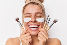 Close Up Portrait Of Happy Woman Keeps Eyes Smiles Toothily Holds Cosmetic Brushes For Applying Makeup Applies Beauty Patches For Reducing Wrinkles Stands Bare Shoulders Isolated Over White Background
