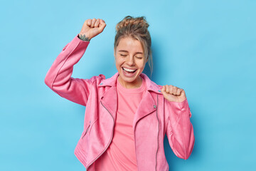 Canvas Print - Happy carefree fair haired woman celebrates victory makes triumph dance clenches fists laughs out joyfully dressed in pink jacket moves against blue background has fun dances on disco party.