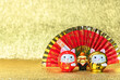 Japanese New Year's Greeting Card depicting a red handheld fan written good luck for a safe house with two cat daruma figurines written good luck against a golden background.