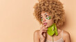 © WHstudio Leushin N - Horizontal shot of tender woman with curly bushy hair keeps hand near mouth focused away applies hydrogel patches under eyes to reduce puffiness holds calla flower poses against beige studio wall