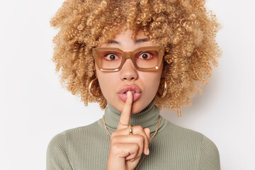 Wall Mural - Mysterious curly haired young woman presses index finger over lips makes silence gesture asks to be quiet wears spectacles and turtleneck isolated over white background. Hush be quiet please